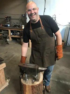Emmanuel smiling behind an anvil in a workshop, in a canvas apron, leather gloves and safety glasses.